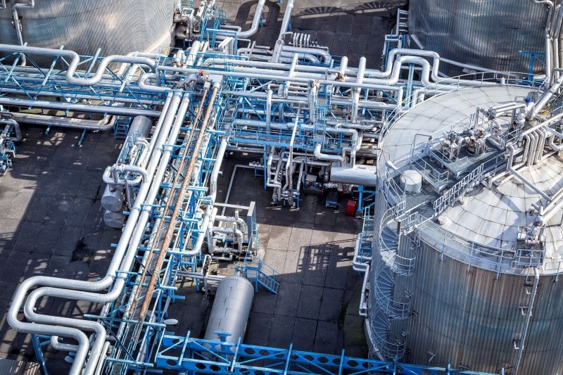 Vista aérea de una refinería industrial que muestra grandes tanques metálicos de almacenamiento y un complejo sistema de tuberías con soportes estructurales azules, válvulas y conductos para el transporte de fluidos.