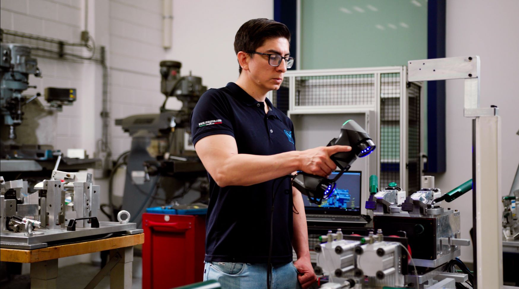 Un hombre con gafas y camisa negra utiliza el escáner HandySCAN 3D para escanear un accesorio mecánico en un taller.