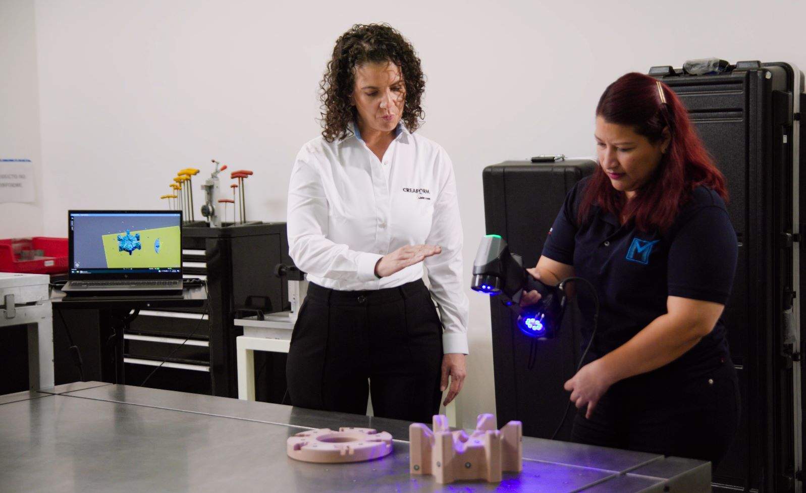 Una experta de Creaform muestra a una empleada de Magove cómo utilizar el escáner HandySCAN 3D para escanear una pieza sobre una mesa en el laboratorio de metrología, mientras el escaneo se muestra en una pantalla al fondo. 