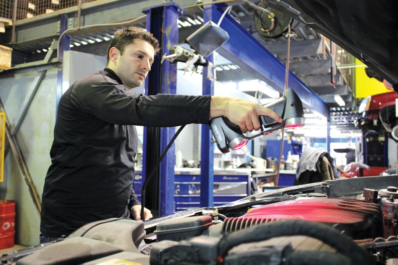 操作人员在工业车间内使用 HandySCAN 3D 手持式 3D 扫描仪扫描汽车发动机,背景中可见各类工具与机械设备