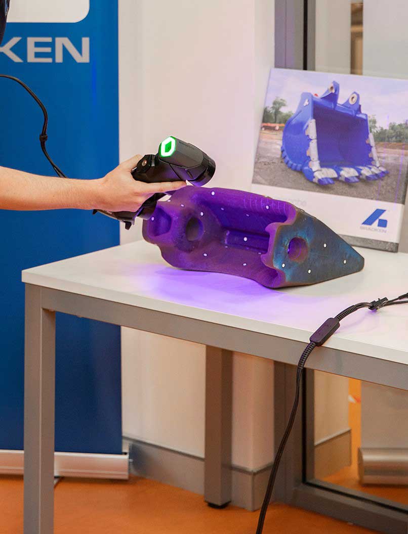 Un operaio Bradken usa uno scanner HandySCAN 3D per scansionare il dente della benna di un escavatore su un tavolo davanti all'immagine di una benna blu.