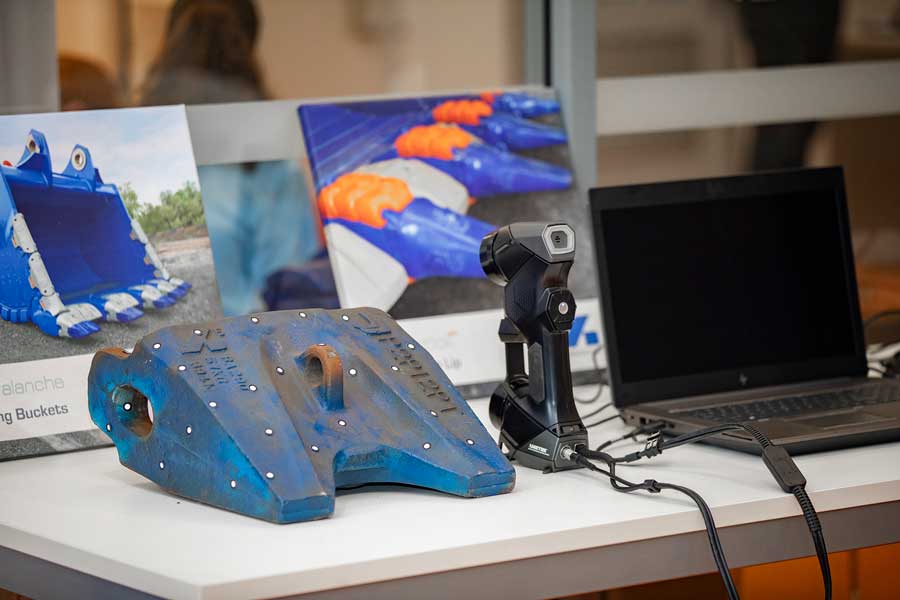 Lo scanner HandySCAN 3D è su un tavolo tra un dente di benna di un escavatore e un portatile nero in front of pictures of the product.