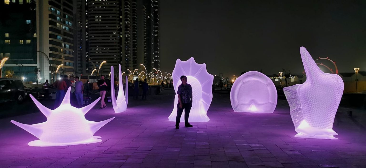 Sculture illuminate di notte con una persona davanti. Le sculture sono immerse nella luce violetta. In secondo piano ci sono dei grattacieli. Alcune finestre sono illuminate