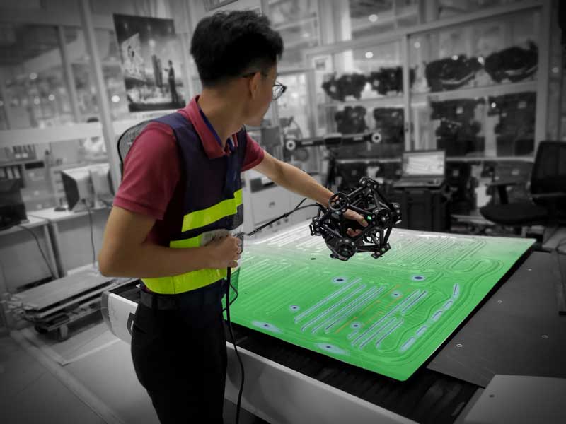 Empleado con chaleco de seguridad utilizando un escáner 3D MetraSCAN para medir una pieza plana de chapa metálica con superposición de mapa de colores.