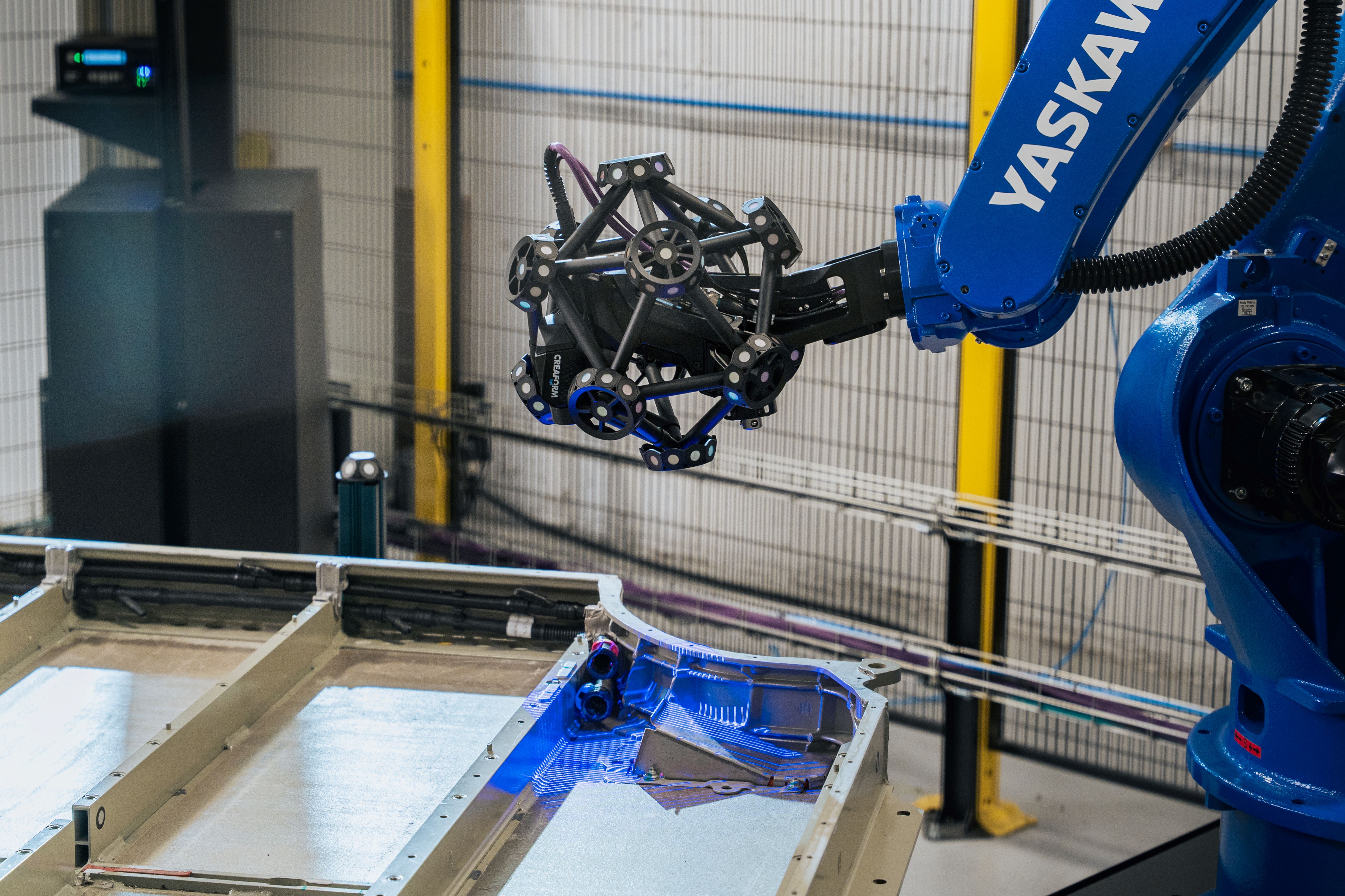 Robot-mounted MetraSCAN 3D-R scanner capturing the geometry of a large metallic component on a workbench, emitting blue light inside an industrial automation cell. 