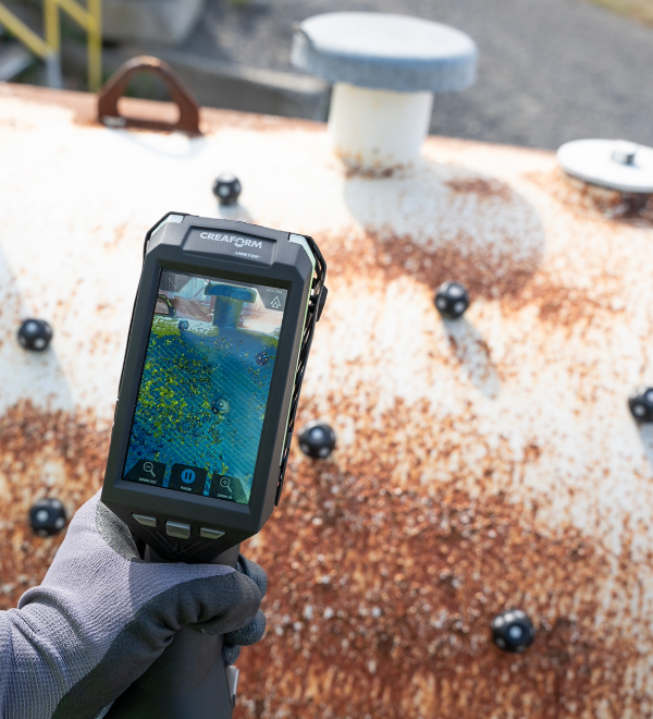 Handheld Creaform scanner capturing 3D measurements of a rusted industrial tank with positioning targets