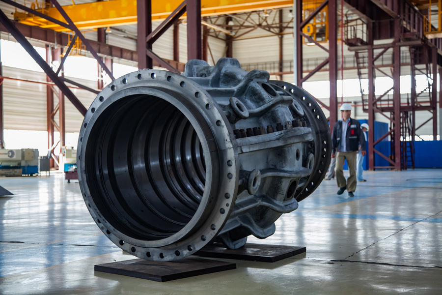 Peça fundida cilíndrica complexa e grande no meio de um hangar, enquanto trabalhadores com capacetes caminham ao fundo